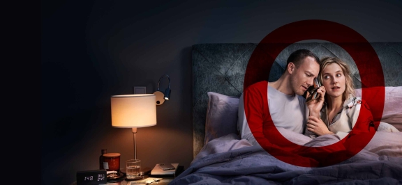 Frau und Mann sitzen aufrecht im Bett. Beide haben Geldbörse am Ohr und hören aufmerksam zu.